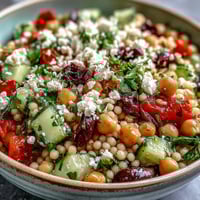 Bright Mediterranean Pearl Couscous salad in a white bowl features toasted couscous, crunchy cucumber, sweet bell pepper, and briny olives.
