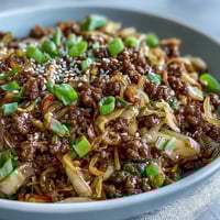 Sizzling ground pork cooks with aromatics for a flavorful Egg Roll in a Bowl garnished with green onions and sesame seeds.